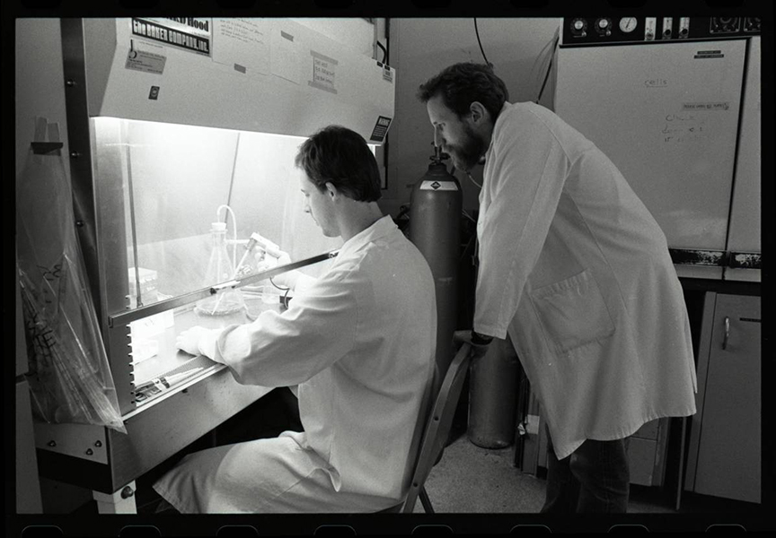 Charles Rice in the lab Black and white photo of two men in front of a lab hood. One is sitting, working in the hood. Charles Rice is standing, watching, to the right.