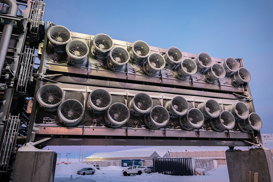 Mammoth direct air capture Photo shows a row of a dozen circular ventilators on each of two metal carbon-dioxide-collector containers with blue sky and a snowy parking lot and buildings in the background.