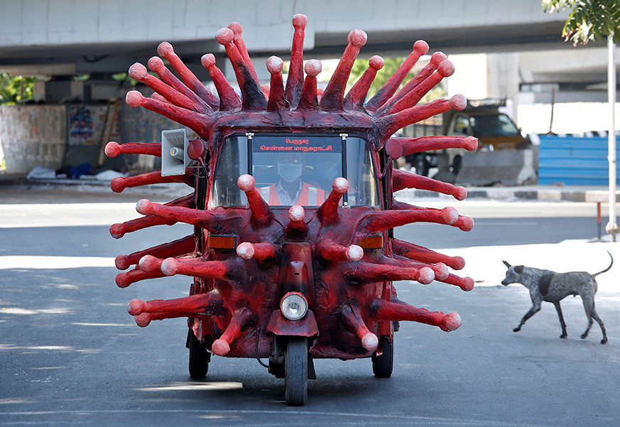 Coronavirus autorickshaw Photograph of an autorickshaw covered with coronavirus-like spikes. A man is driving it down a street; a dog looks on.