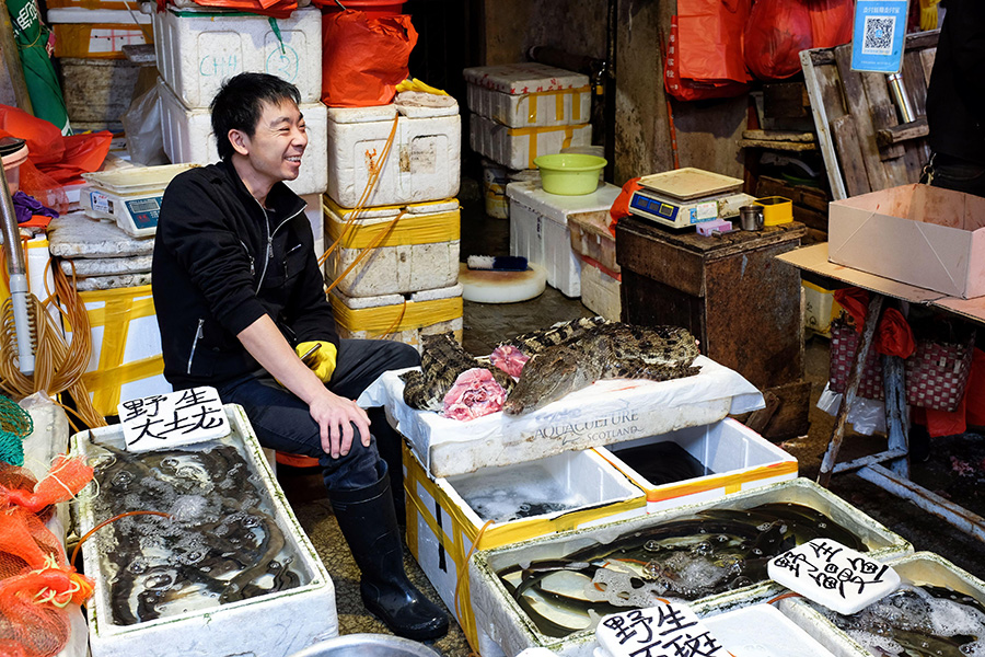 Crocodile meat for sale A man in black clothing sits smiling, surrounded by containers and displays of different types of raw flesh.