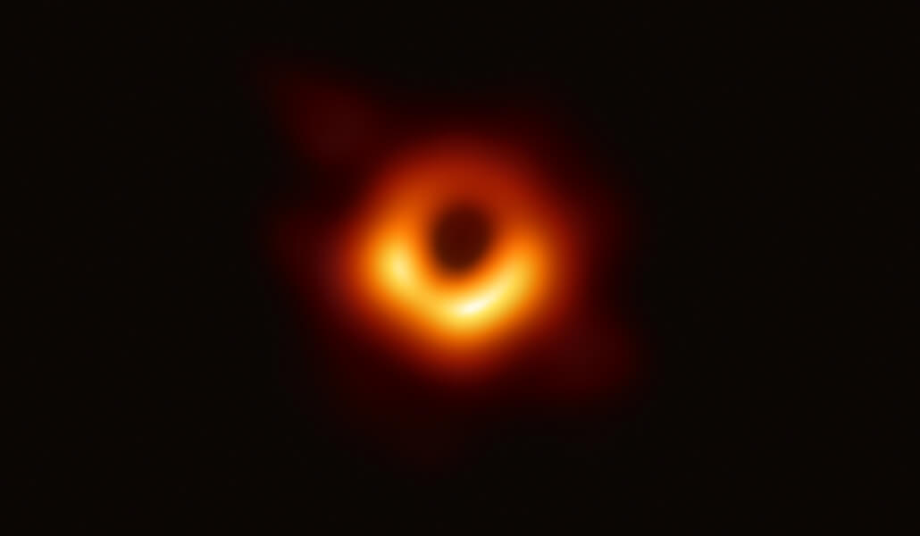 Black hole Image shows a dark shadow ringed in bright red and yellow formed as light bends in the intense gravity around a black hole.