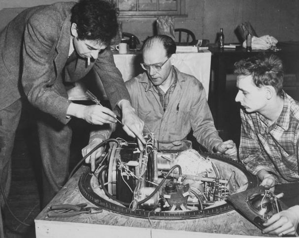 Cosmic ray detector Old photo shows man in suit standing, cigarette in mouth, while adjusting machinery on an instrument while a seated technician and younger man look on.