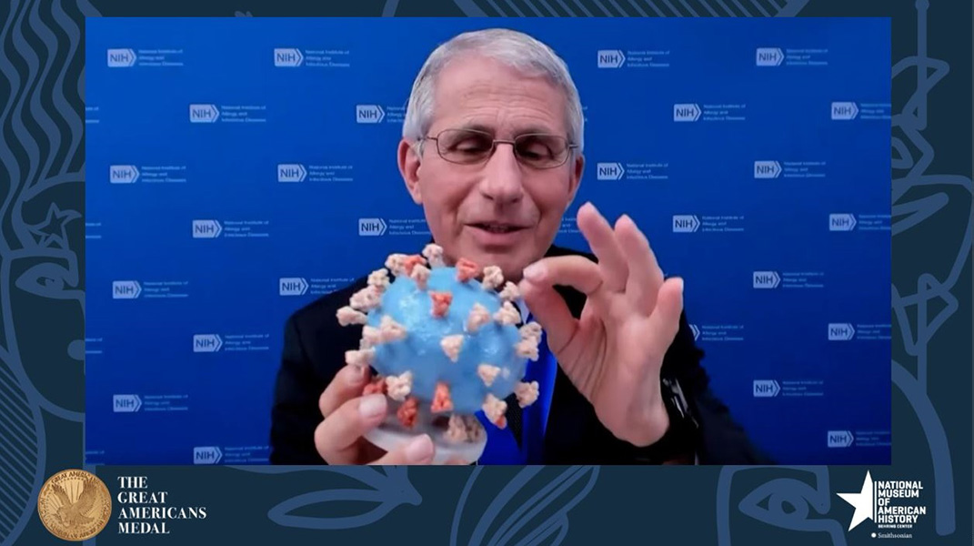 Fauci coronavirus model Photograph of Anthony Fauci holding an educational coronavirus model: It is a round blue blob with red spikes protruding from it.