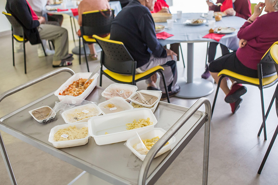 Bland, beige foods Photo of food in cartons on a tray; nearby are tables with nursing home residents sitting around them. The foods are bland shades of brown and beige.