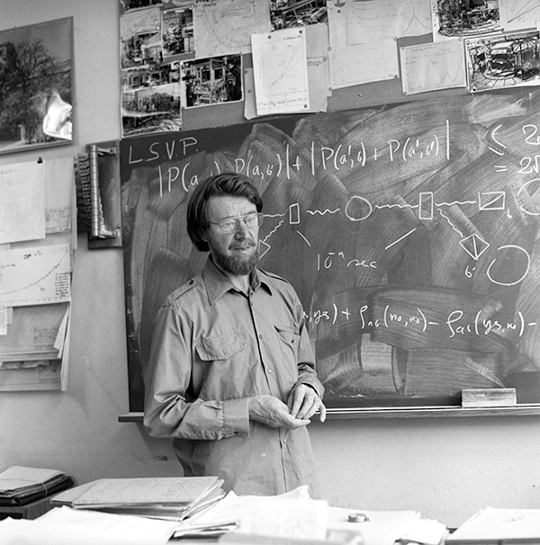 John Stewart Bell Historic photo shows John Bell standing in front of a chalkboard with equations and drawings on it, with papers piled on a table in front of him and tacked to the wall around the chalkboard.