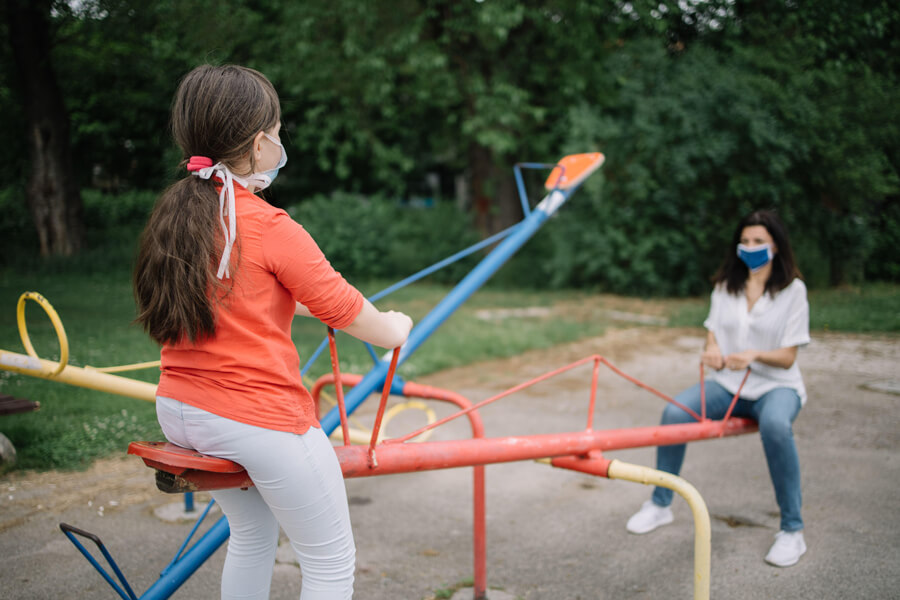 Playground Photo shows a girl and a woman wearing protective masks playing on a see-saw at playground.