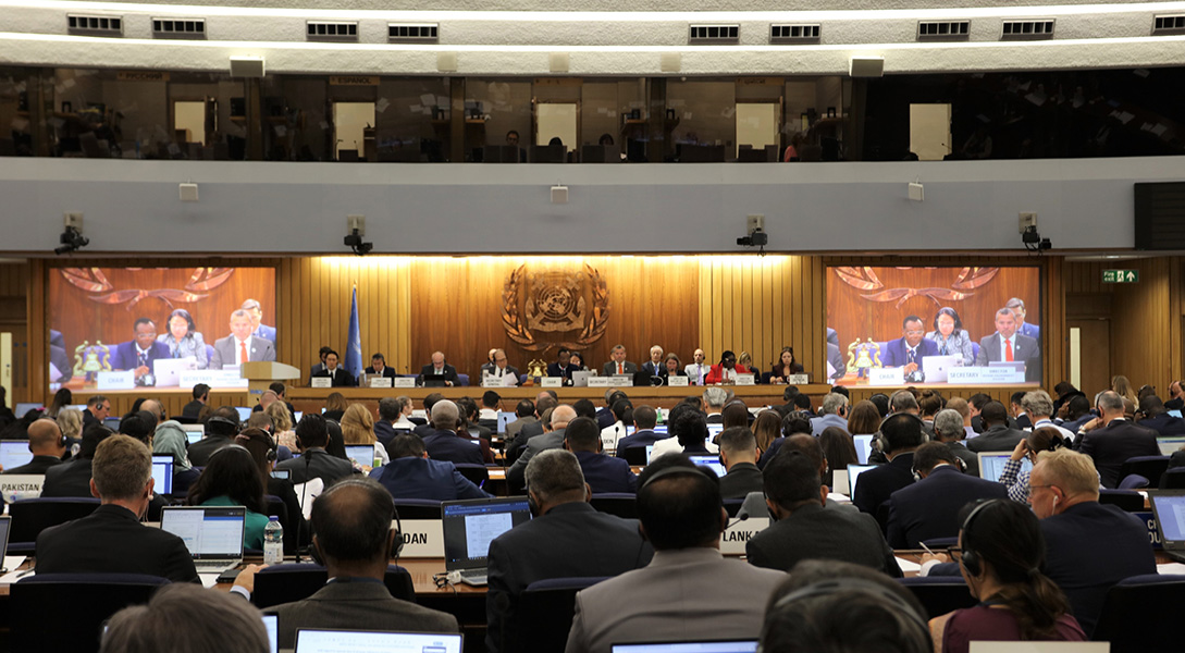 IMO meeting Photograph of a meeting taking place in a large room, with panelists up on stage and a packed audience watching and takes notes on laptops. The IMO logo is up on the wall behind the stage.