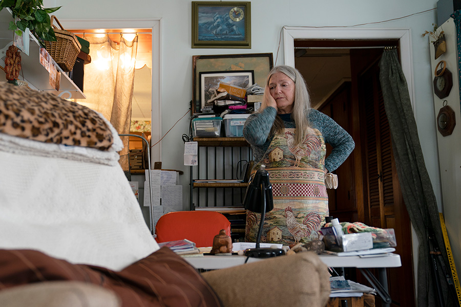 Long Covid Photograph of a woman with gray hair wearing an apron. She is standing in front of a desk in her home and has one hand held up to her face.