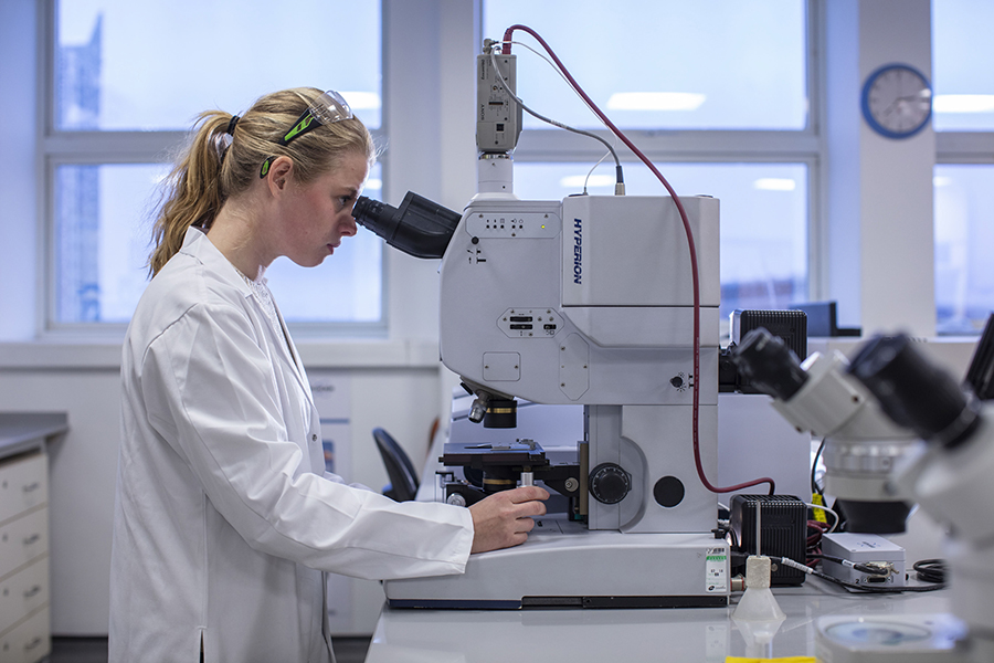 Imogen Napper microsope Photograph of Imogen Napper wearing white lab coat. She is looking through the eyepiece of a microscope. There are windows behind her.