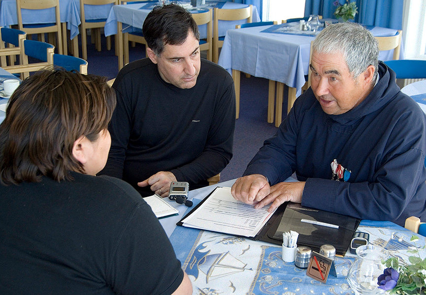 Greenlandic elder Three people are seated at a table. One of them, an older man, is talking and referring to a notebook, while the other two listen intently.