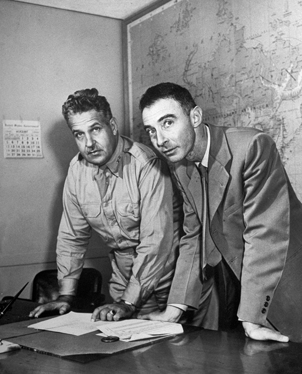 Manhattan Project leaders A black-and-white photo of two men, one in a military uniform and the other in a suit, stand before a desk, with a map behind them.