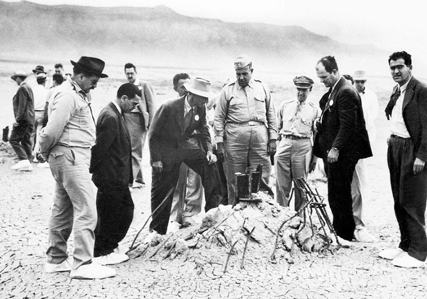 Trinity test site A black and white photo shows a small group of men in suits and uniforms surrounding a mound of dirt with bent steel rebar poking up through it, against a desert backdrop.
