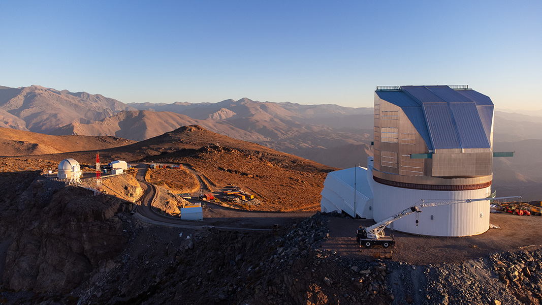 Vera C. Rubin Observatory A sunset view of Rubin Observatory atop its Chilean desert mountain site; mountain ridges recede into the distance under a blue sky.