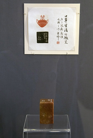 Smile Behind the Mask Photograph of a museum exhibit. A carved seal is standing on a small glass table. Above it is a printed image with Chinese calligraphy.