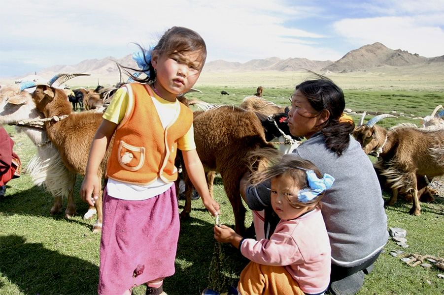 Tuvan family Photograph of a woman squatting on the ground, with two children in front and goats in the background.