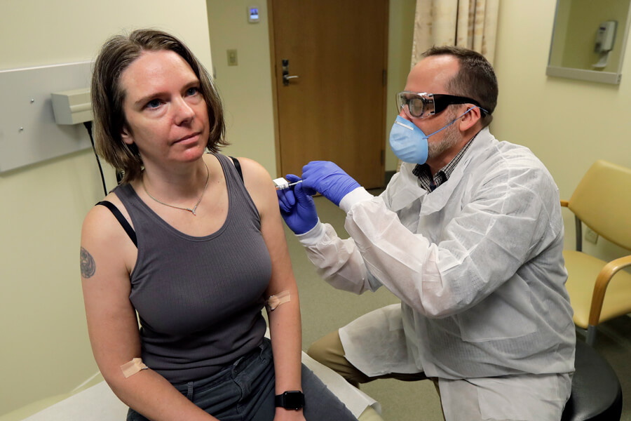 Volunteer in vaccine trial Photograph of a woman in a tank top sitting on a bench receiving a shot in the arm. A man wearing a mask, goggles, gloves and a white coat is administering the shot.